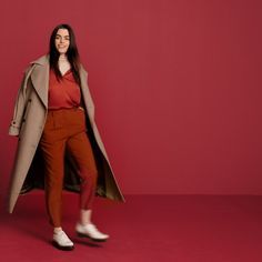 a woman standing in front of a red background wearing a trench coat and orange pants