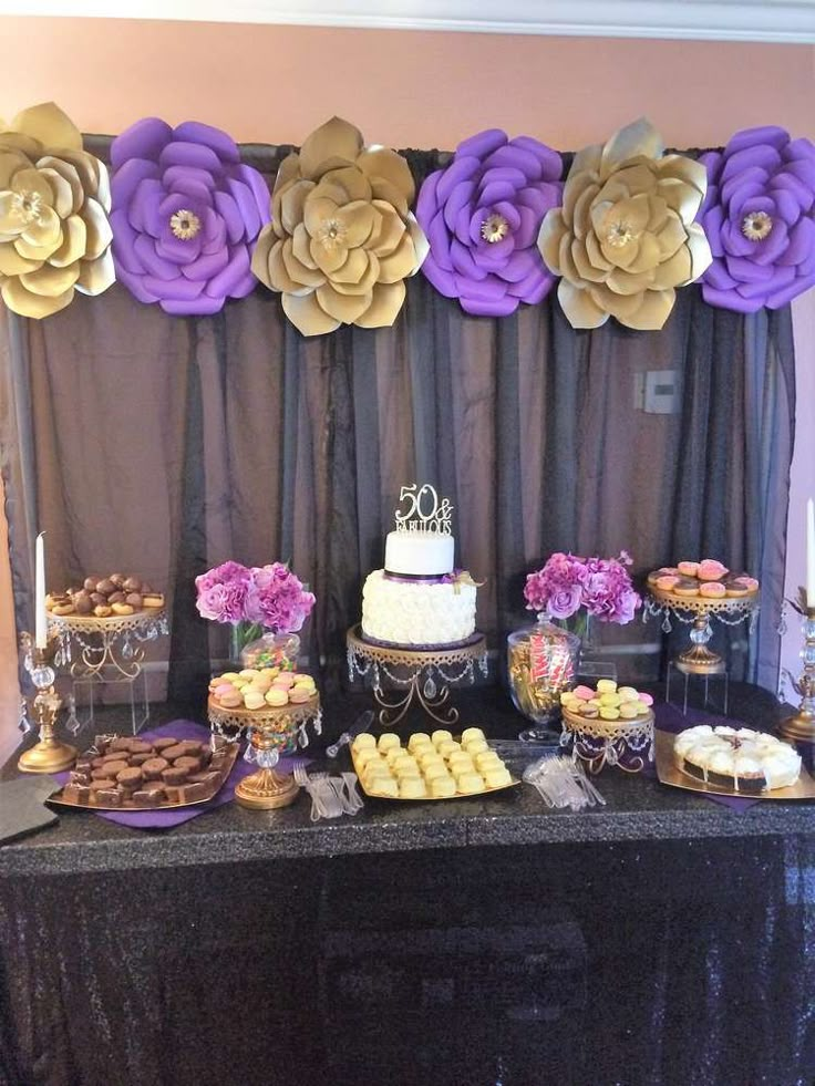 a table topped with cakes and desserts covered in purple paper flower decorations on top of a black table cloth