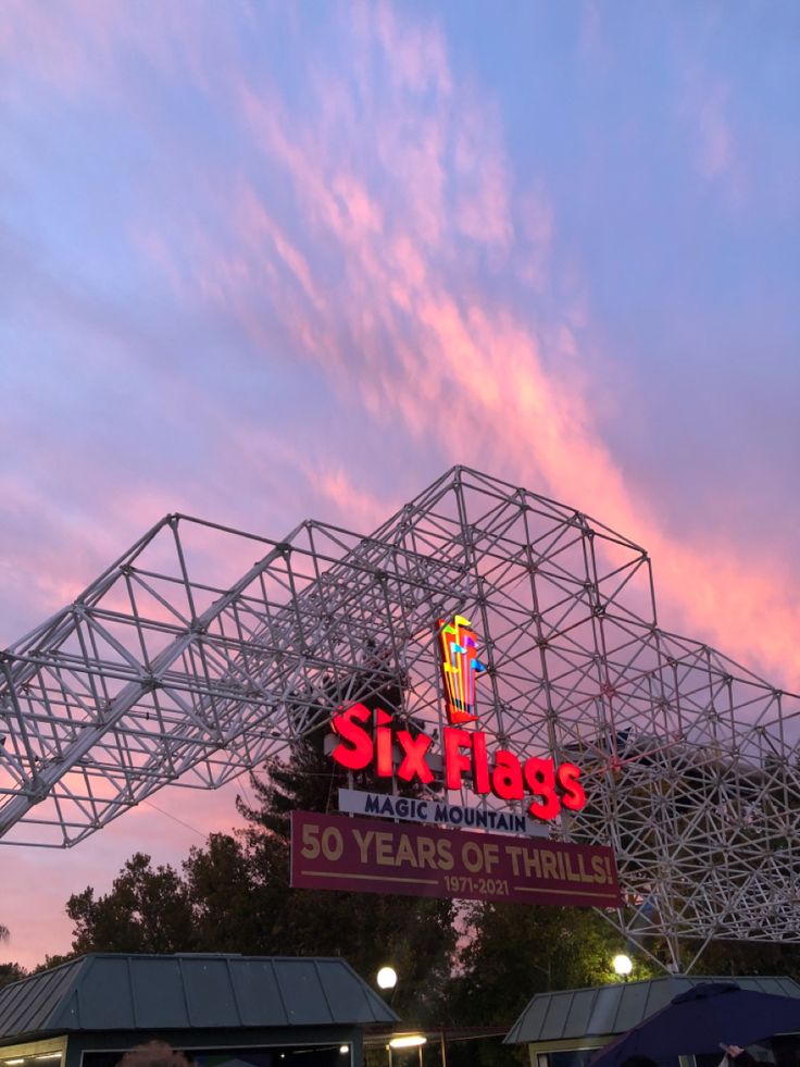 the six flags sign is lit up at dusk with pink clouds in the sky behind it