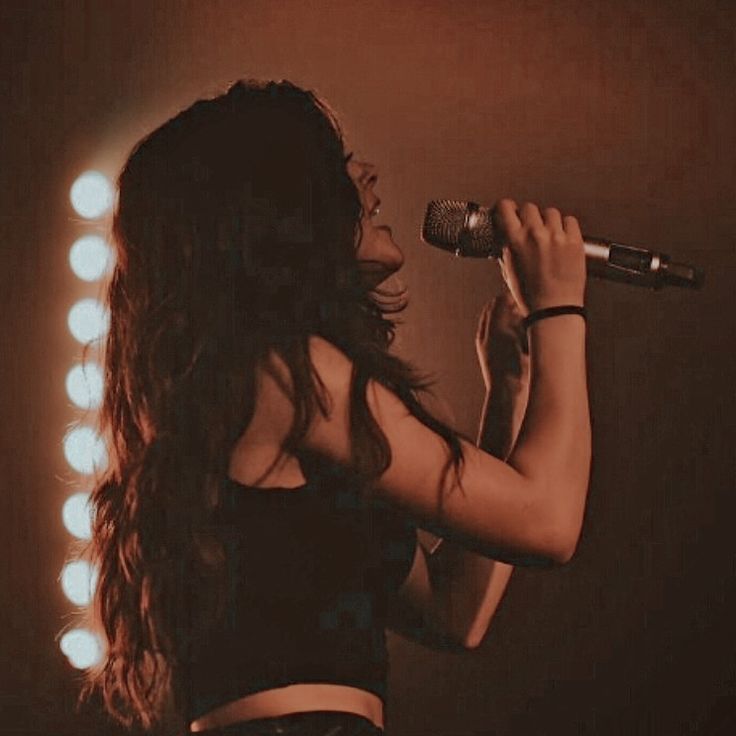 a woman singing into a microphone in front of a mirror with lights on the wall behind her