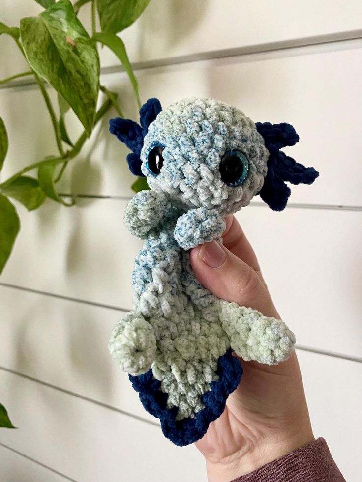 a hand holding a small crocheted stuffed animal in front of a plant with green leaves