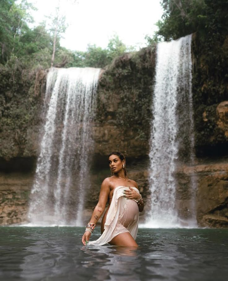 a woman standing in the water next to a waterfall with her arms wrapped around her waist