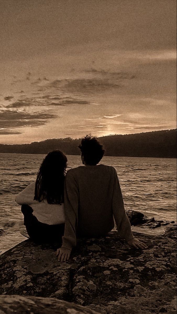 two people sitting on rocks looking out over the water at sunset or sunrise with clouds in the sky