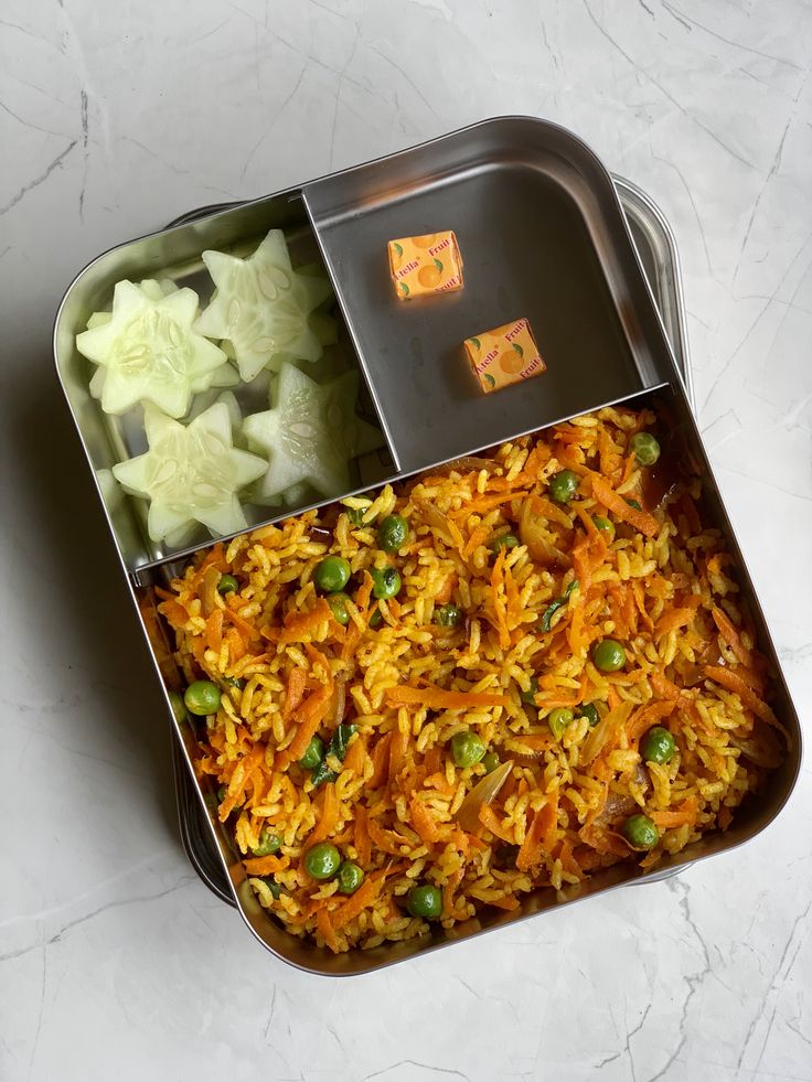 a metal container filled with rice and vegetables on top of a white countertop next to sliced cucumbers