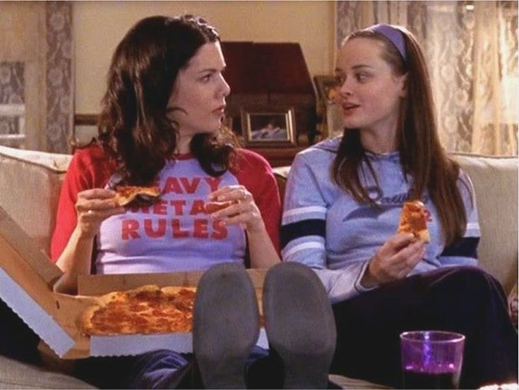 two women sitting on a couch eating pizza and talking to each other in front of them