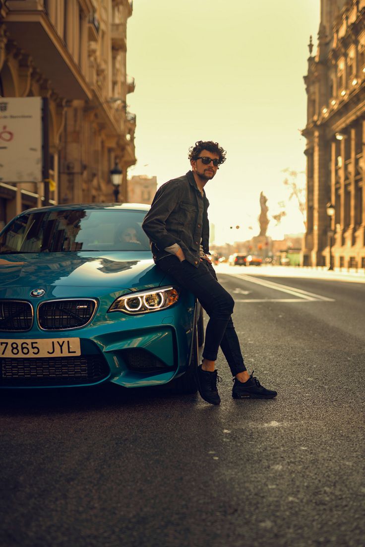 a man sitting on the hood of a blue car in front of a street with tall buildings