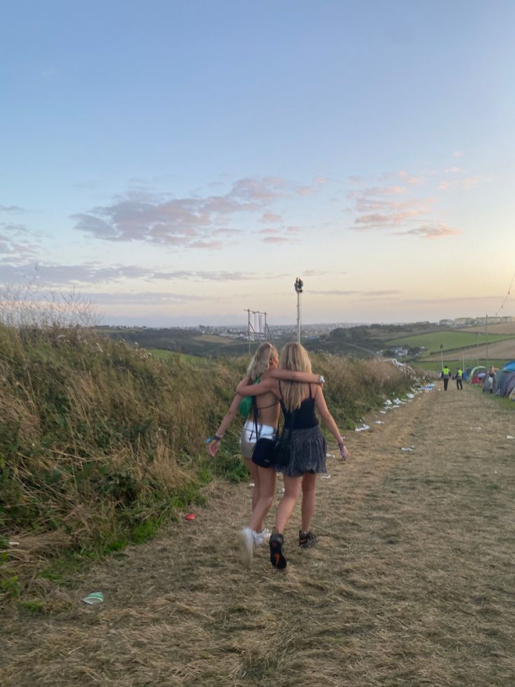 two young women walking down a dirt road next to grass and tents in the distance