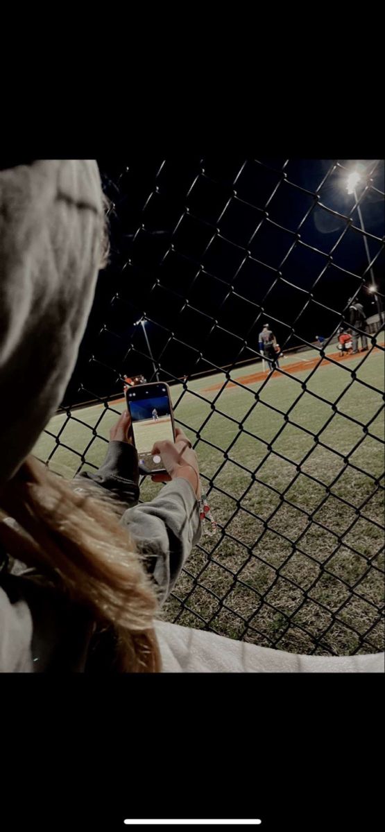 a woman taking a photo with her cell phone in front of a baseball field at night