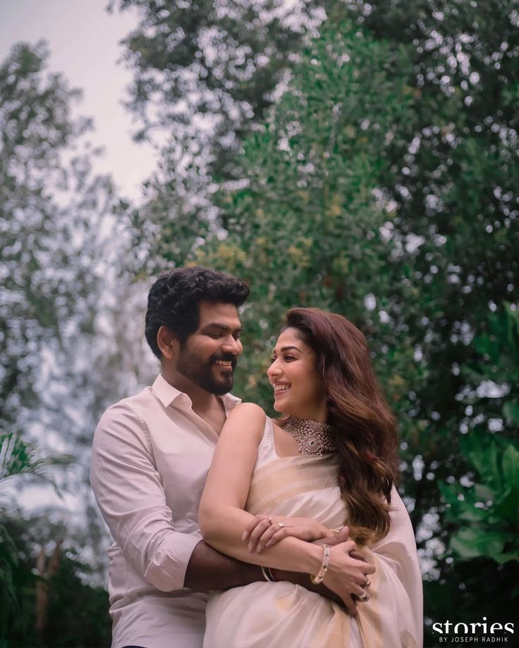 a man and woman standing next to each other in front of trees with their arms around each other