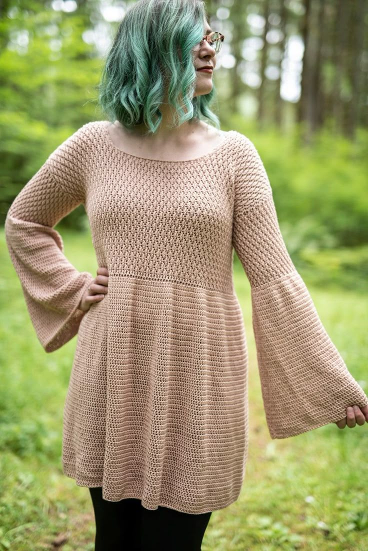 a woman with green hair is standing in the woods wearing a brown sweater and black leggings