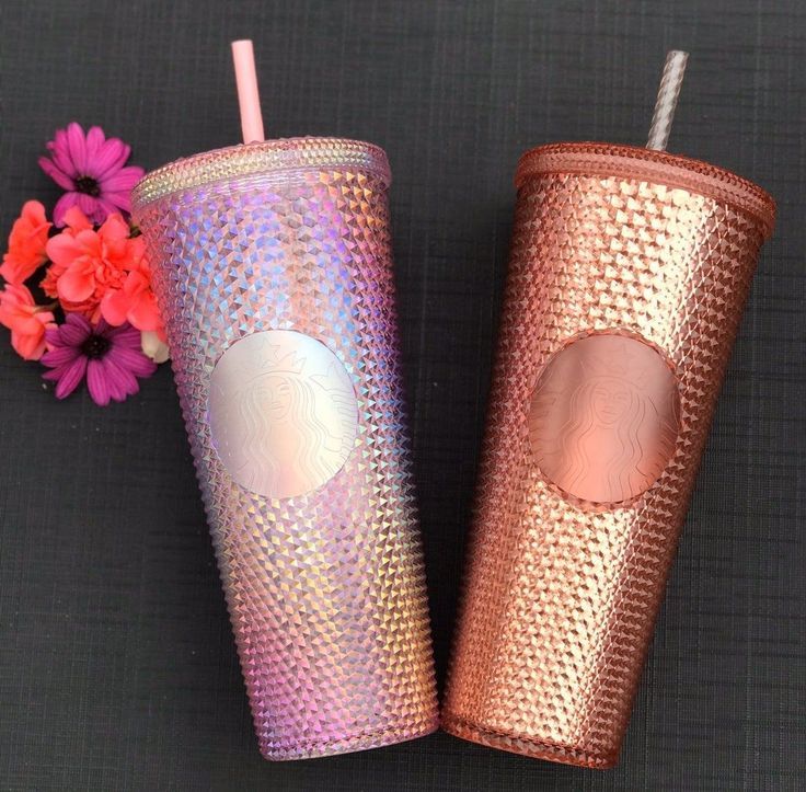 two pink and purple starbucks cups with straws next to flowers on a black surface