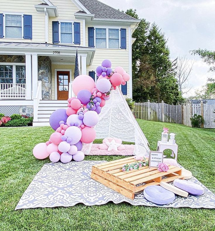 a pink and purple balloon arch is on the lawn next to a picnic table in front of a large white house