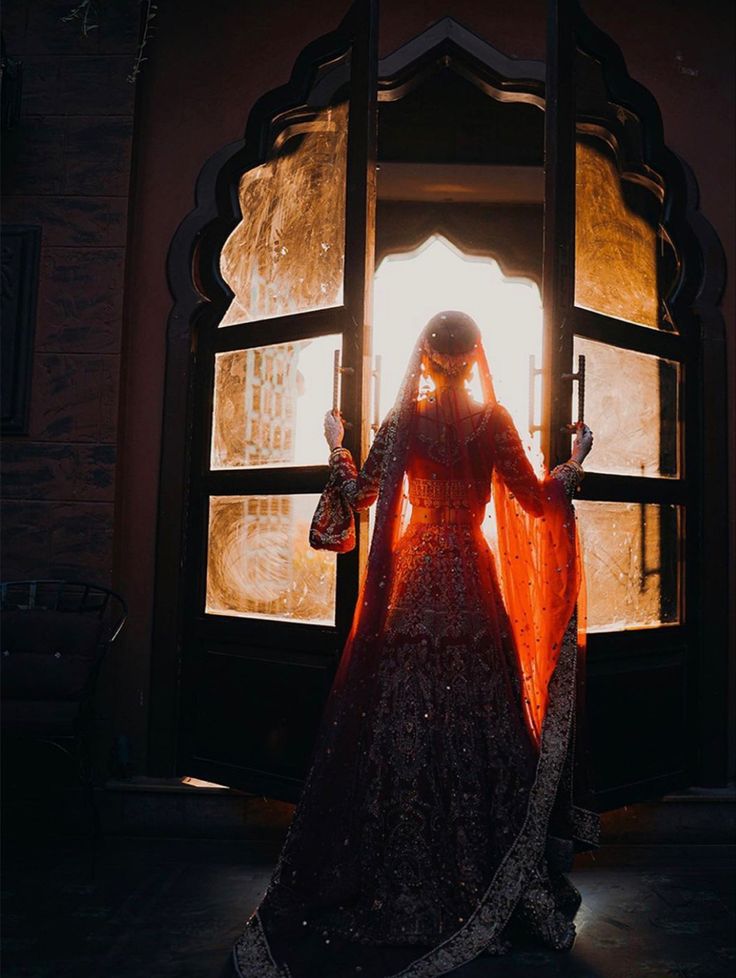 a woman standing in front of an open window wearing a red and black outfit with long veil