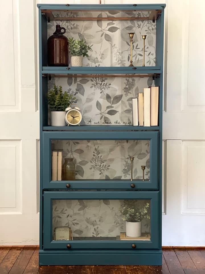 a blue bookcase with books and plants on it in front of a wallpapered background