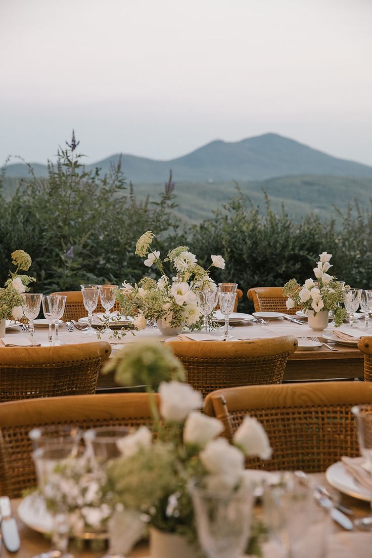 an outdoor table set with white flowers and place settings in front of the mountain range