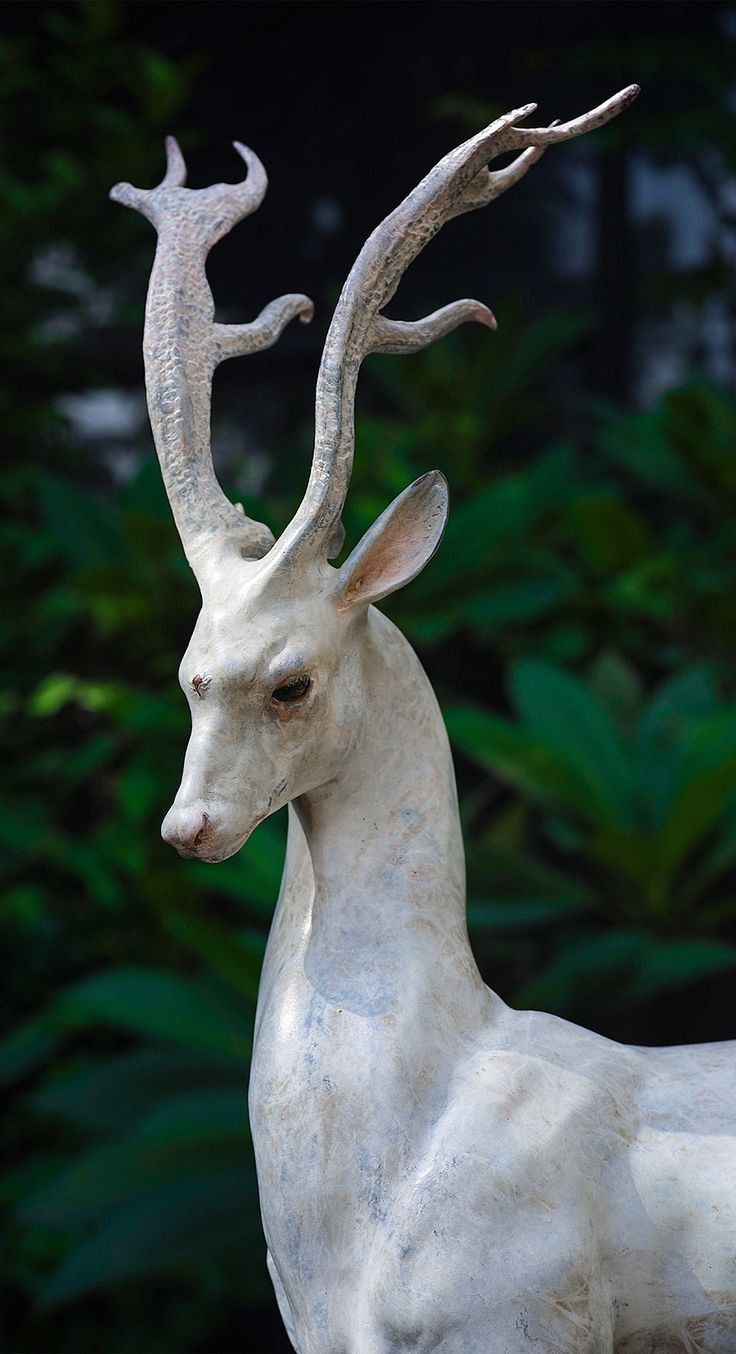 a statue of a white deer with horns on it's head, standing in front of some bushes