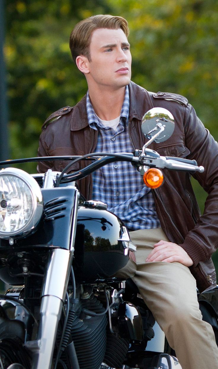 a man sitting on top of a motorcycle wearing a leather jacket and khaki pants