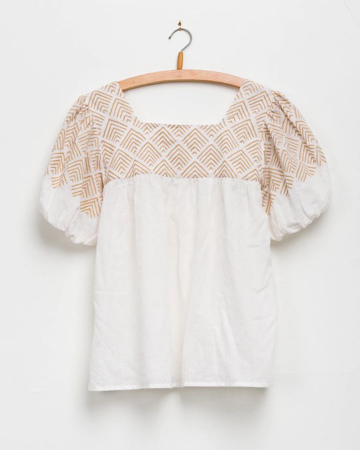 a white top with gold print on the chest and shoulders, hanging from a wooden hanger