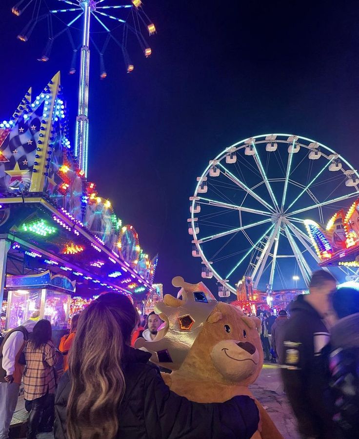 people walking around an amusement park at night with rides and ferris wheel in the background