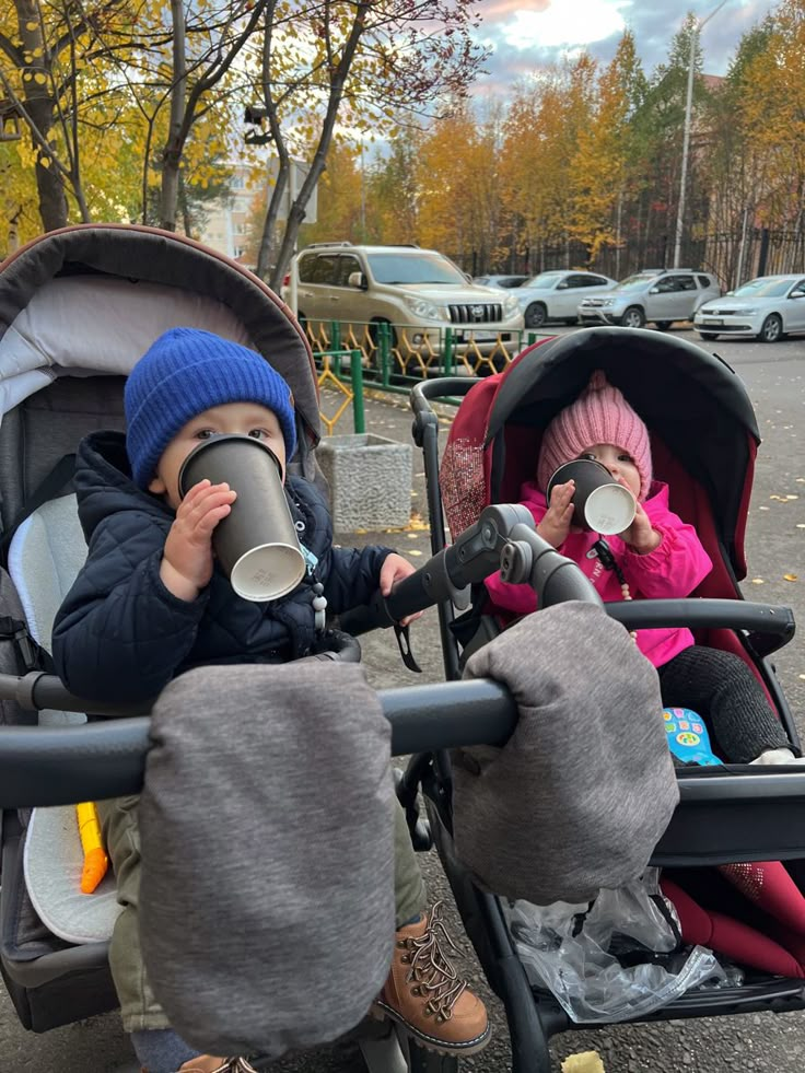 two small children sitting in strollers on the side of the road, one holding a coffee cup