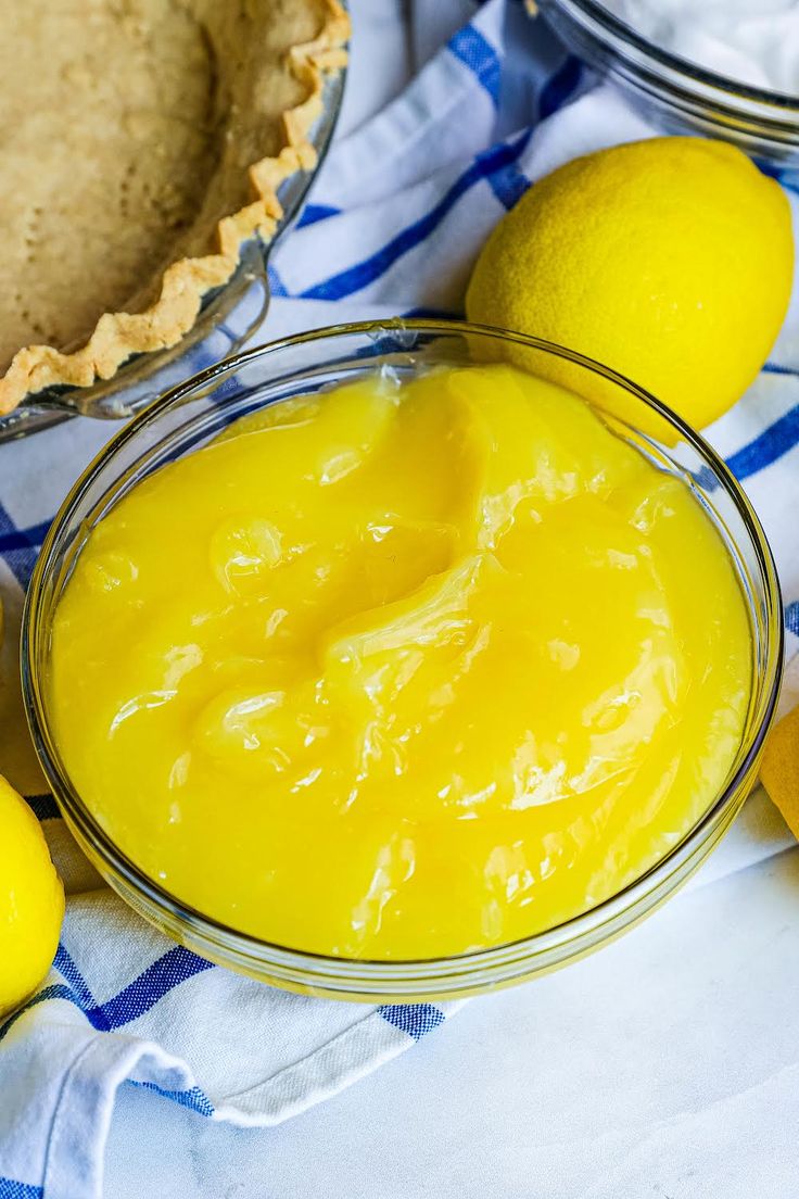 a glass bowl filled with yellow liquid next to two lemons and a pie crust