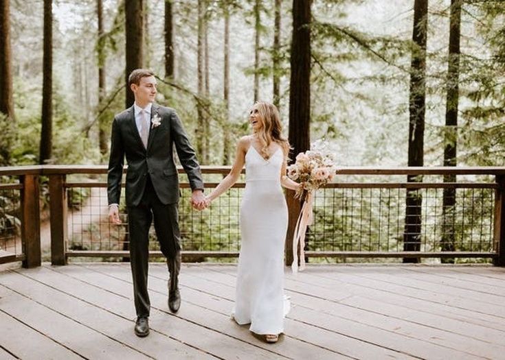 a bride and groom holding hands while walking on a deck in the middle of a forest