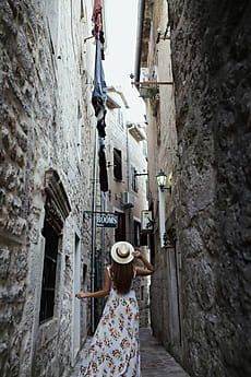 a woman in a dress and hat walks down an alleyway between two stone buildings