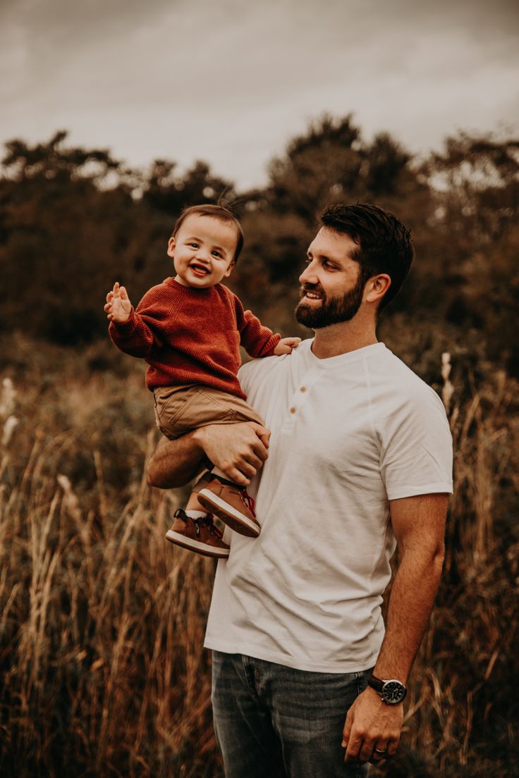 a man holding a toddler in his arms and smiling at the camera while standing in tall grass