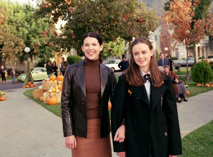 two women standing next to each other in front of pumpkins on the side walk