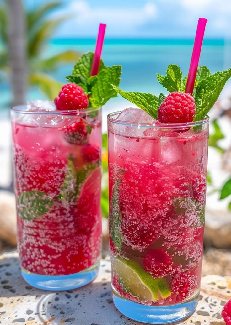 two glasses filled with raspberry mojitas on a table near the ocean