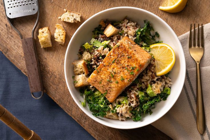 a white bowl filled with rice, broccoli and fish next to lemon wedges