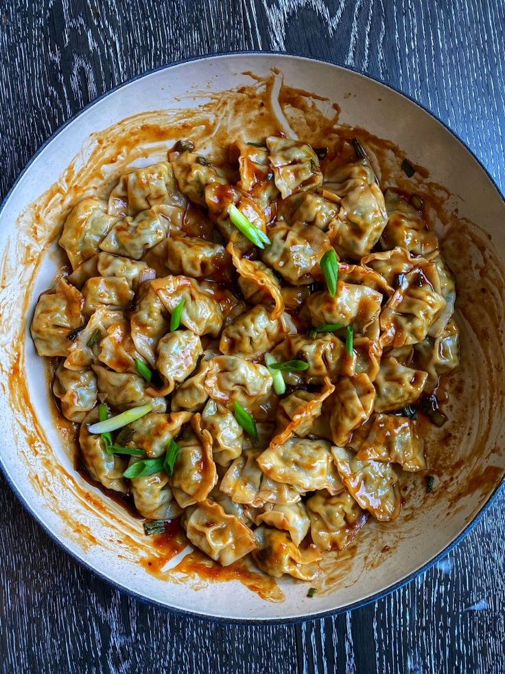 a white bowl filled with dumplings covered in sauce and green onions on top of a wooden table