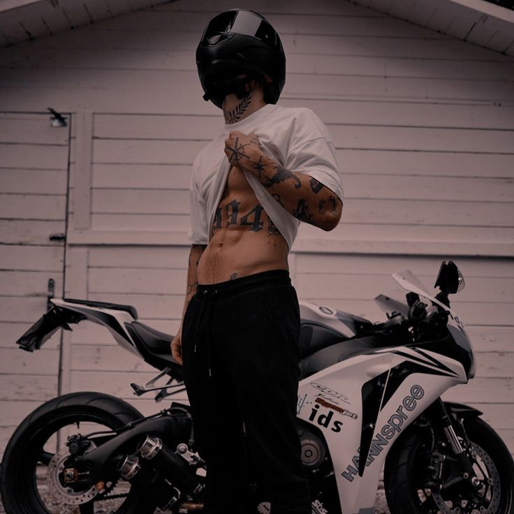 a shirtless man standing next to a motorcycle in front of a garage with his hand on his chest