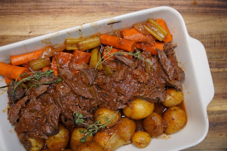 a white dish filled with meat and potatoes on top of a wooden table next to carrots