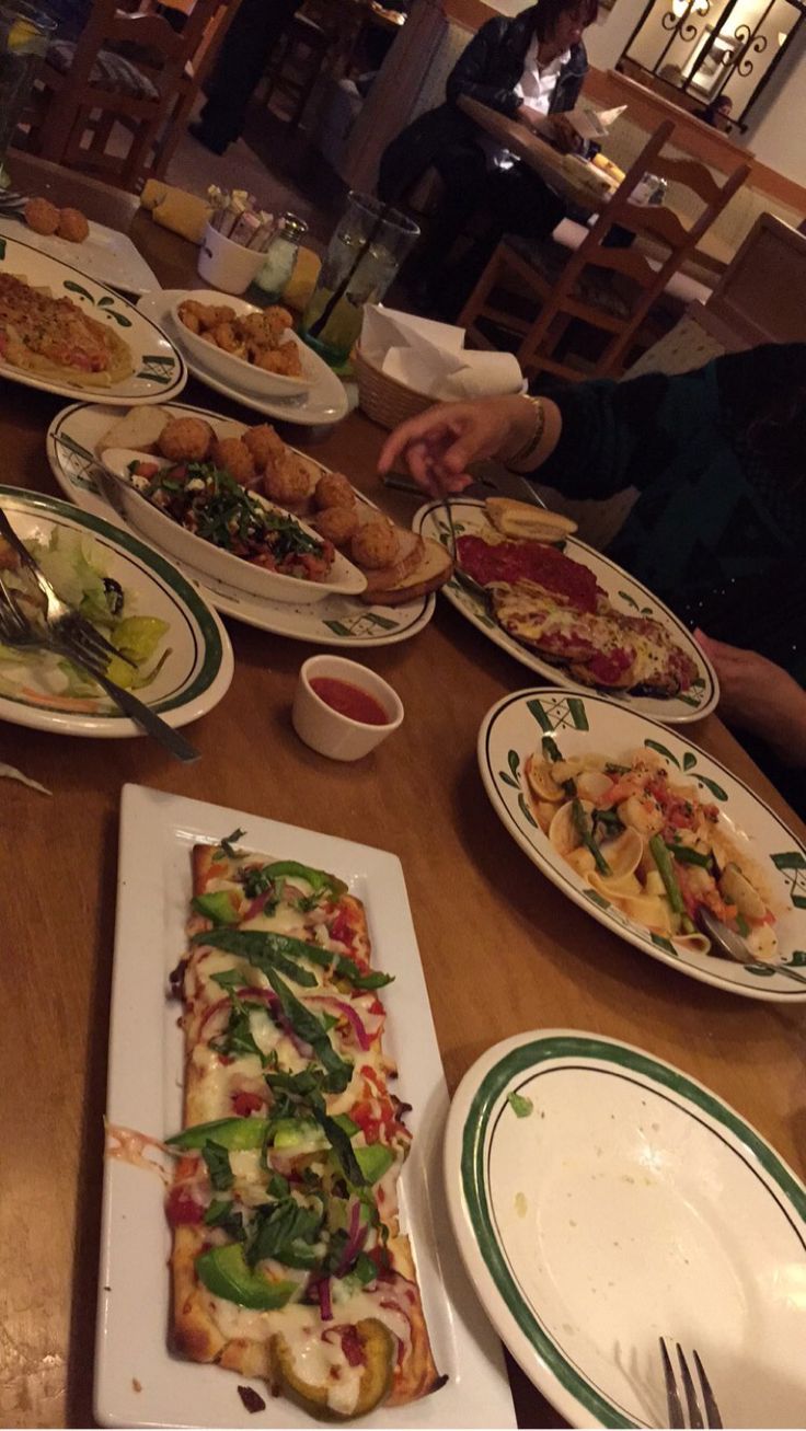 people sitting at a table with many plates of food on it, including pizza and salad