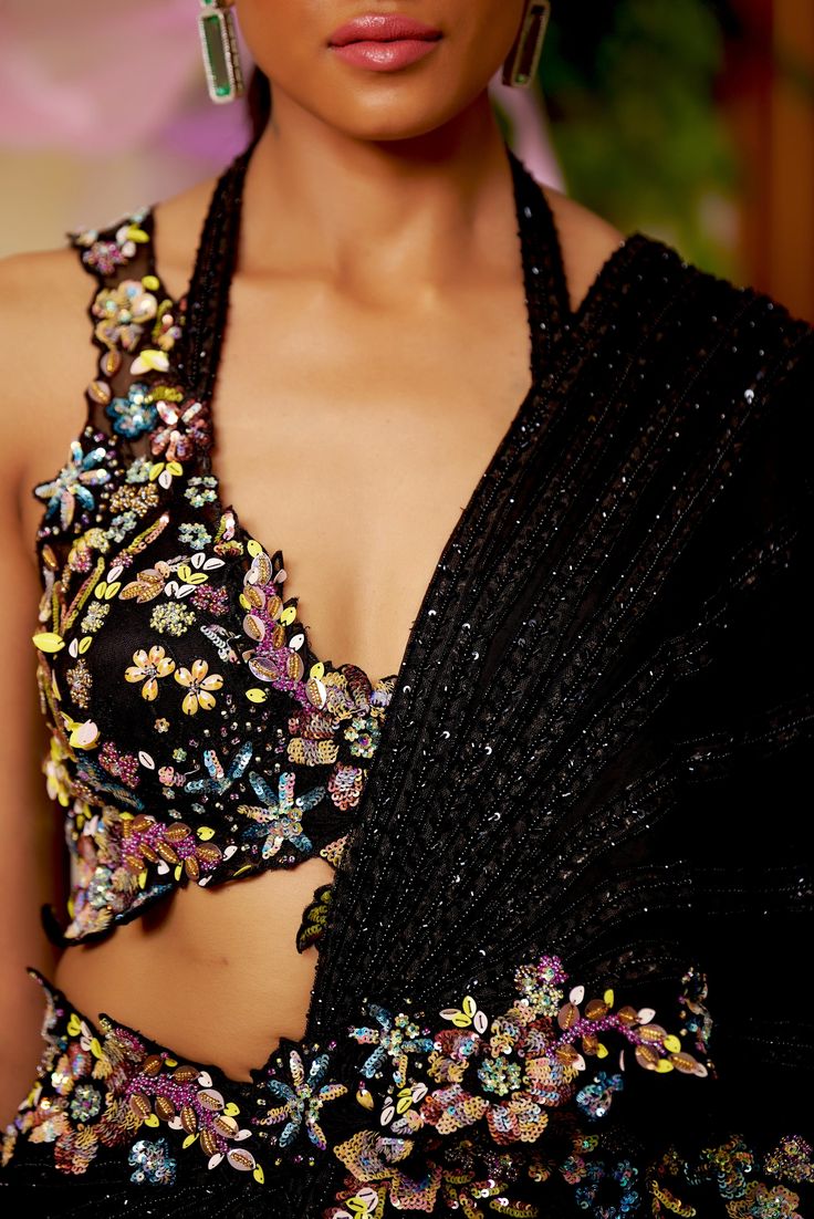 Black is your new summer romantics to go all glam for any intimate soiree of the season. This pre-draped saree highlighted with the lines of black bead-work all around comes with a dazzling floral blouse accentuated with embroidered multicolor bead-work and cut-work. The floral bead embroidery also highlights the waistline and goes on the pallu’s borders. Sleeveless blouse with a deep neck and cutwork detailing at the edges. Hook at the back of the blouse. The silk satin pre-draped saree is acce Saree Edging Ideas, Black Saree Blouse, After Wedding Dress, Indian Wardrobe, Draping Styles, Reception Outfits, Indian Room, Fancy Stuff, Draped Saree