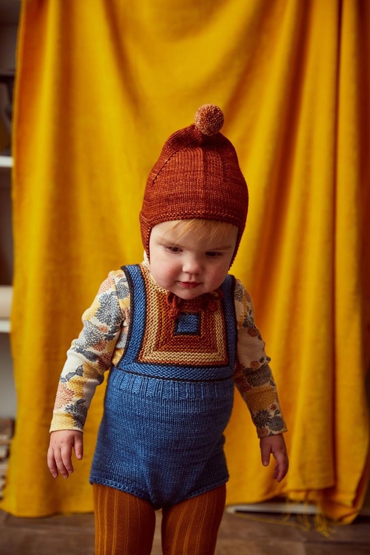 a small child in overalls and a knitted hat is standing on the floor