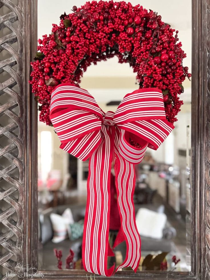 a red and white wreath with a bow hanging from the front door, on top of a mirror