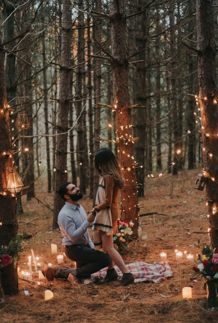 a man kneeling down next to a woman on a blanket in the woods surrounded by candles