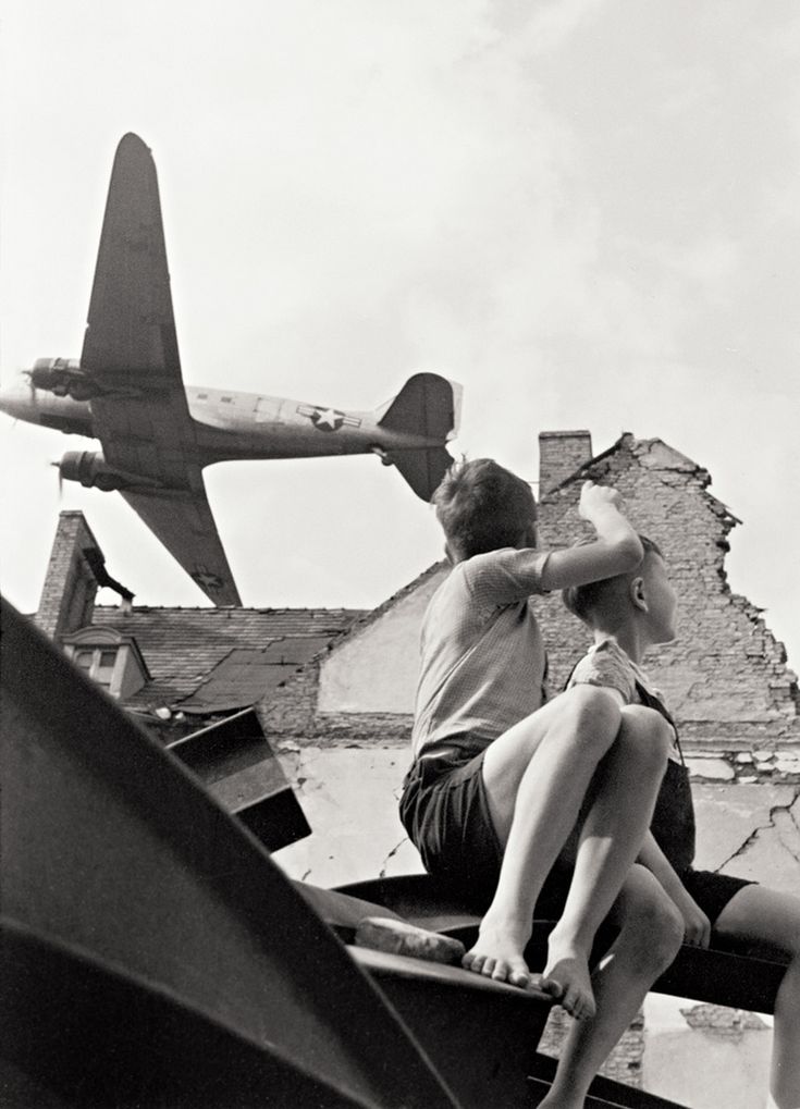 black and white photograph of two people sitting on the back of a truck with an airplane in the background