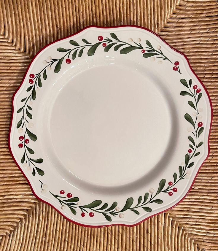 an empty white plate with red trim and green leaves on it sitting on a wicker surface