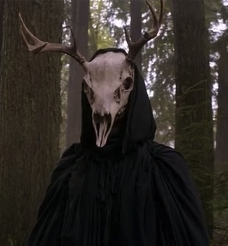 a woman in a black dress with an antelope mask on her head standing next to trees