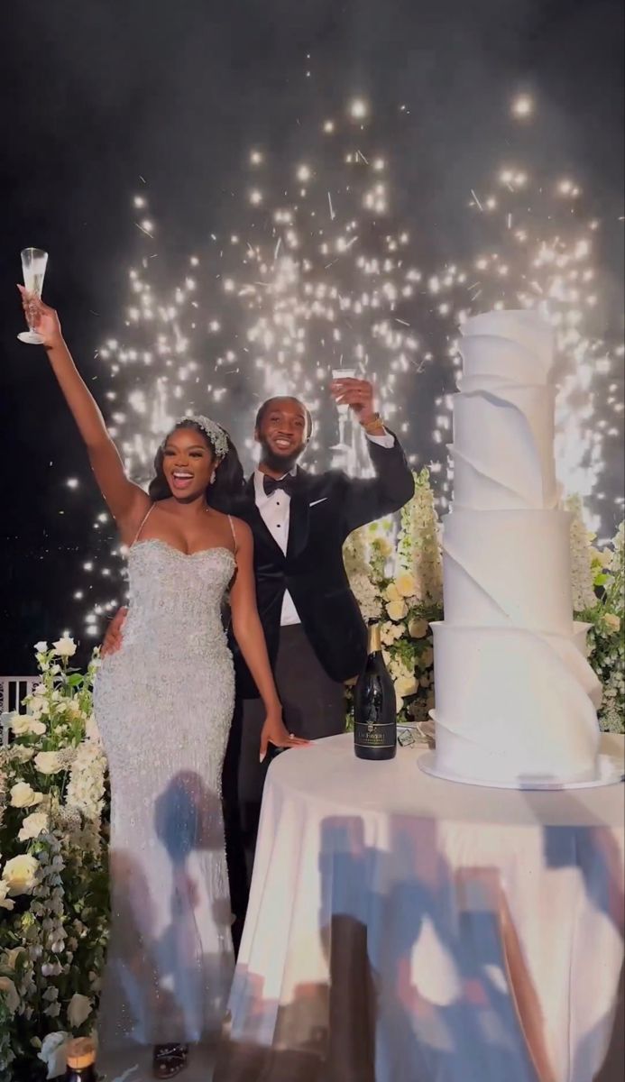 a newly married couple standing next to a wedding cake with fireworks in the sky behind them