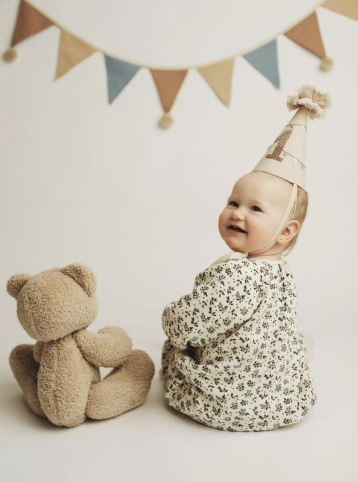 a baby sitting next to a teddy bear wearing a party hat