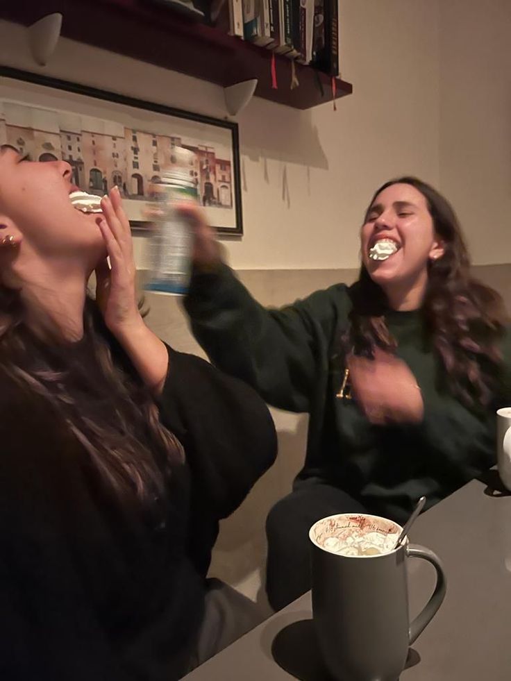 two women sitting at a table eating cake and drinking coffee while one woman holds her mouth open