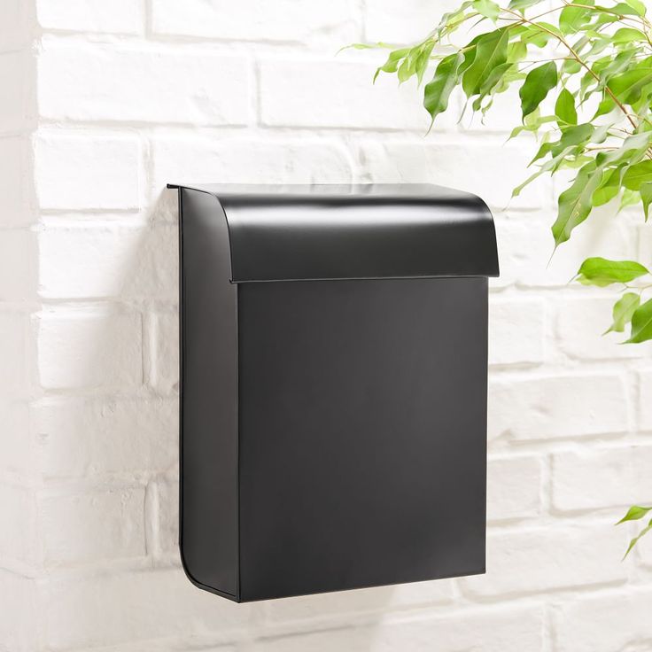 a black mailbox mounted to the side of a white brick wall next to a green plant