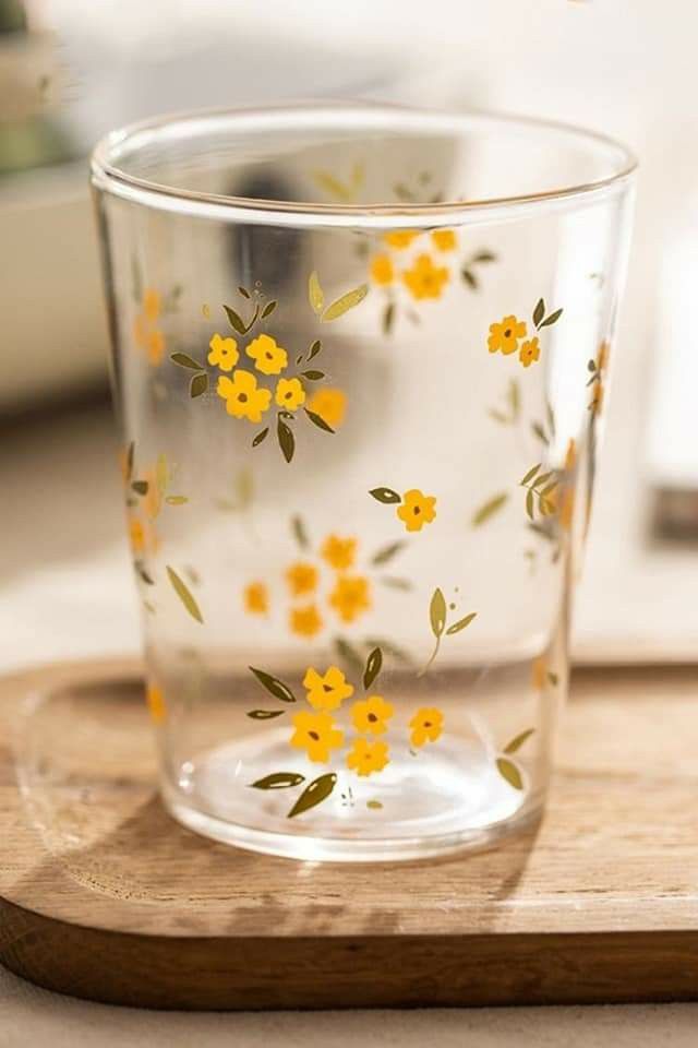 a glass with yellow flowers painted on it sitting on a wooden tray next to a window