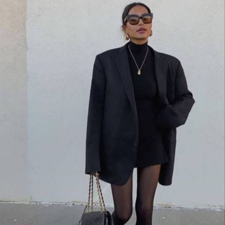 a woman is standing in front of a white wall holding a handbag and wearing black tights