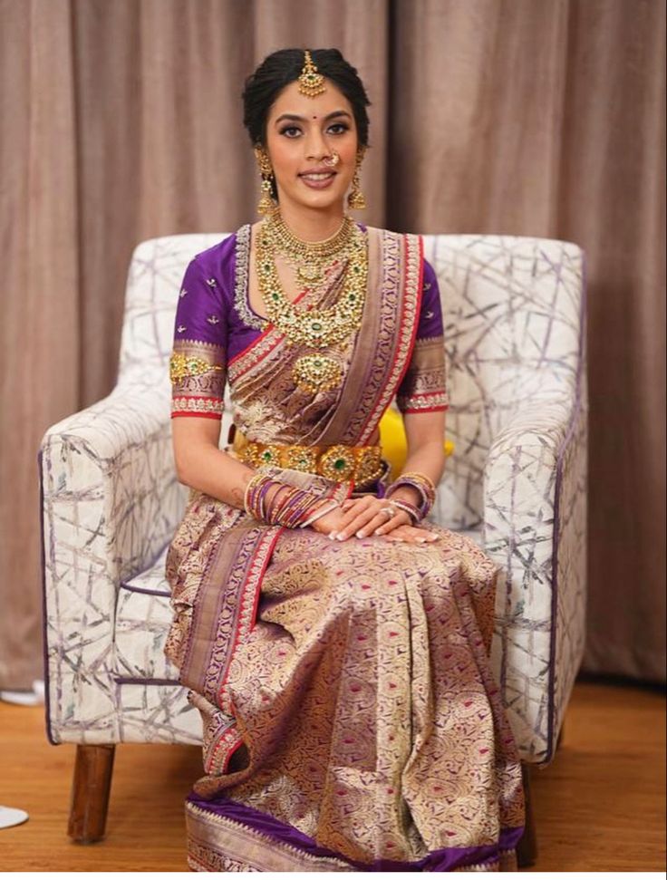 a woman sitting in a chair wearing a purple and gold sari with jewelry on her neck