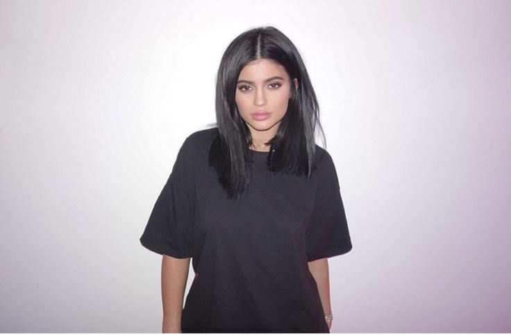 a woman with long black hair standing in front of a white wall wearing a black t - shirt